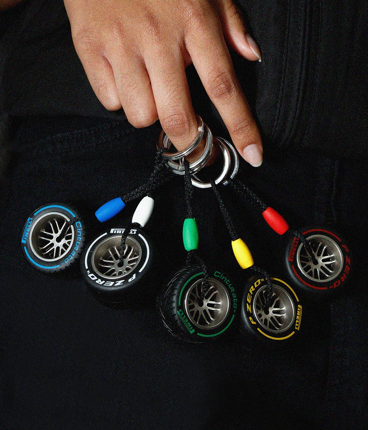 Pirelli Tyre Keyrings with tyre designs hanging from rings, held by a hand against a black background. F1® merchandise.